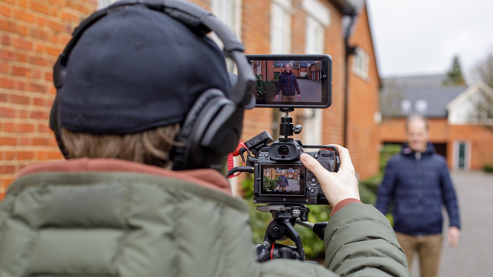 A person behind a camera filming a man outdoors for an online videography course, with a focus on the camera's monitor showing the live shot.