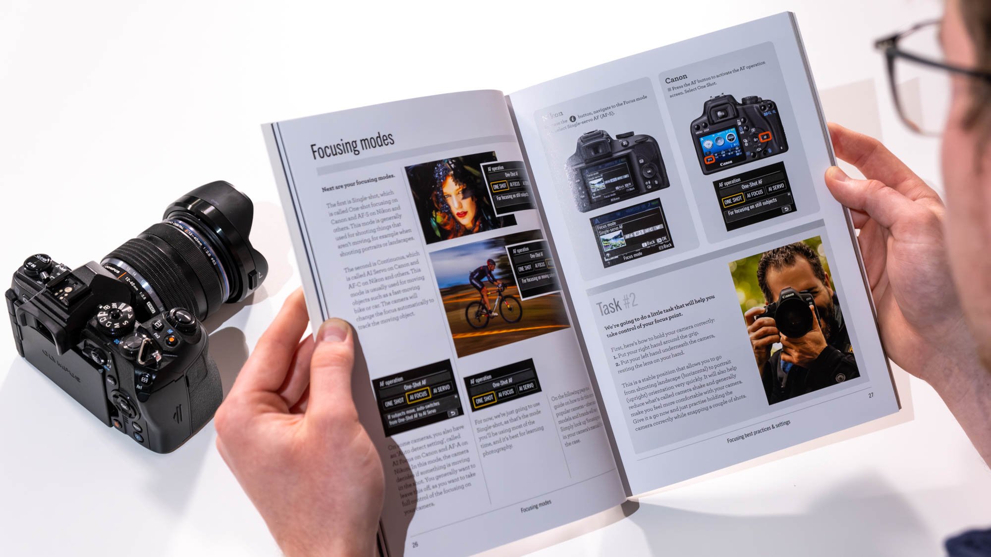 Person reading a photography book about camera focusing modes next to a mirrorless camera.