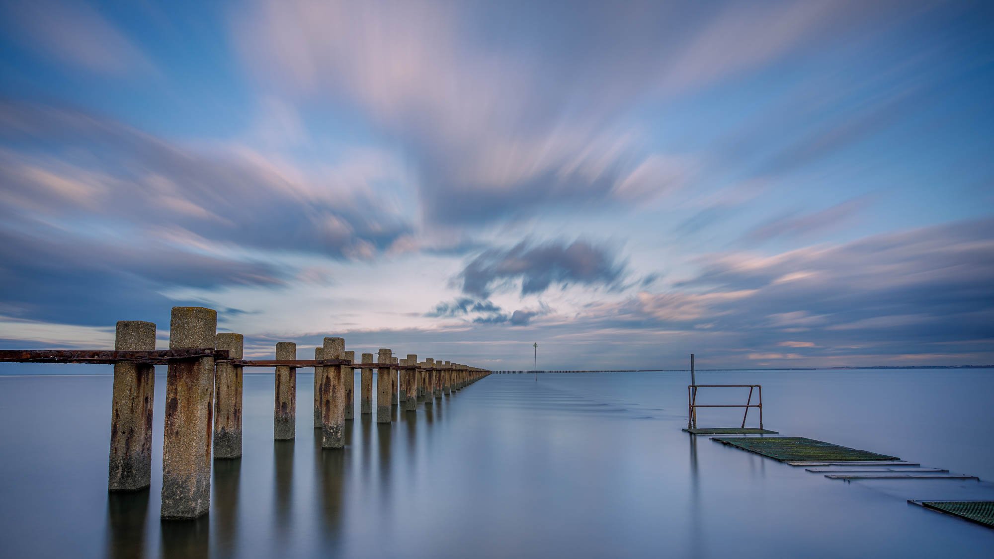 Long Exposure of calm water and streaky clouds over a weathered pier extending into the sea.