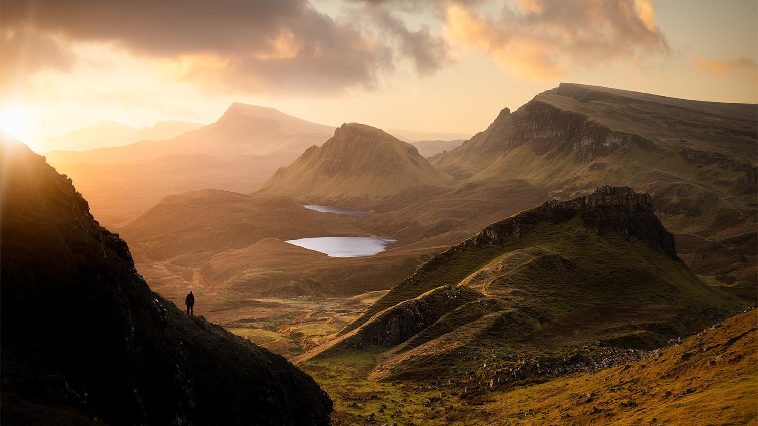 Golden light illuminates a person in a vast, dramatic mountain landscape with lakes.