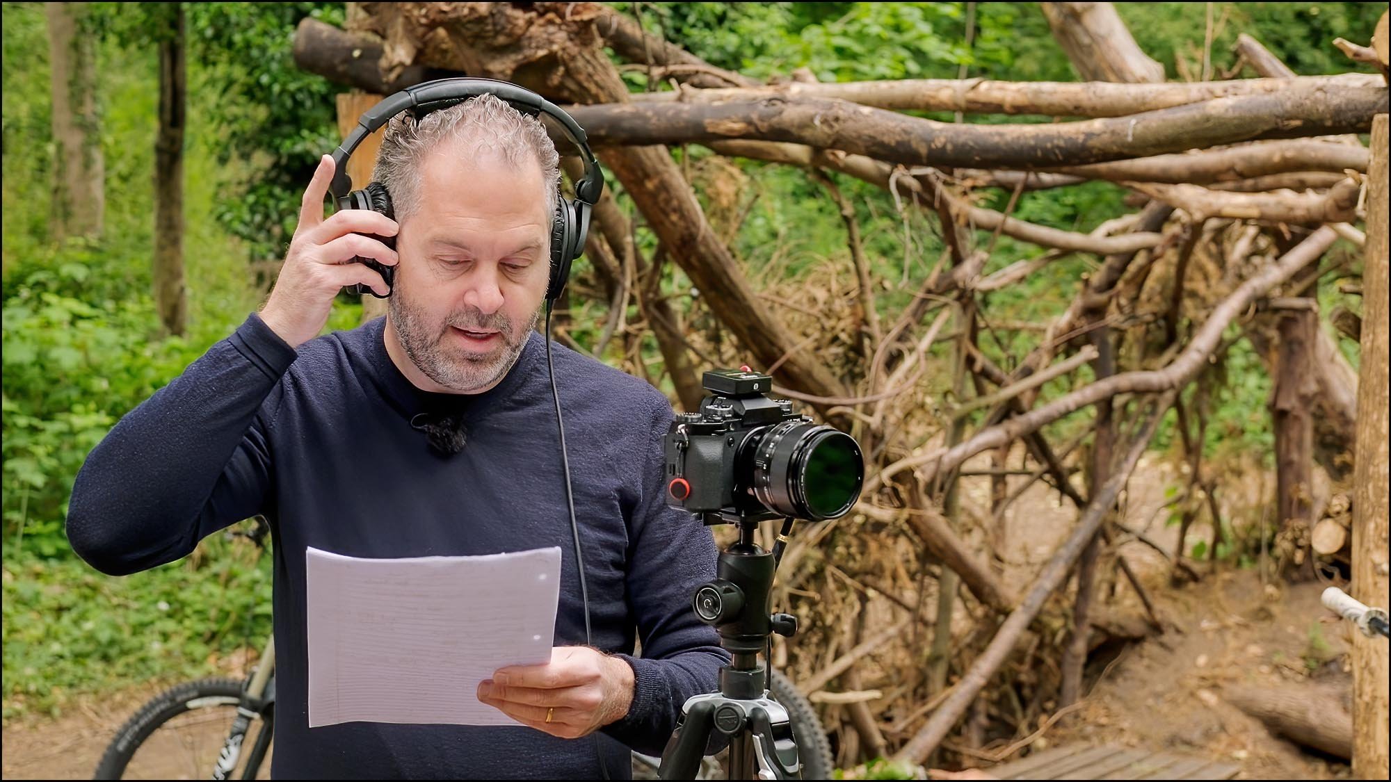 Videography instructor reviewing a script while monitoring audio with headphones beside a camera on a tripod during a planning lesson