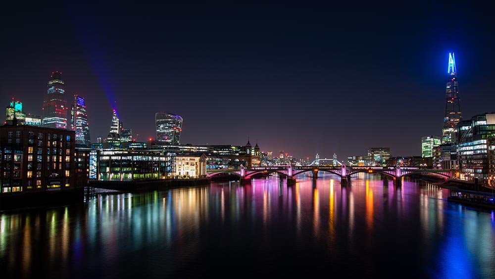 London skyline at night reflecting in the Thames, illuminated bridges and buildings.